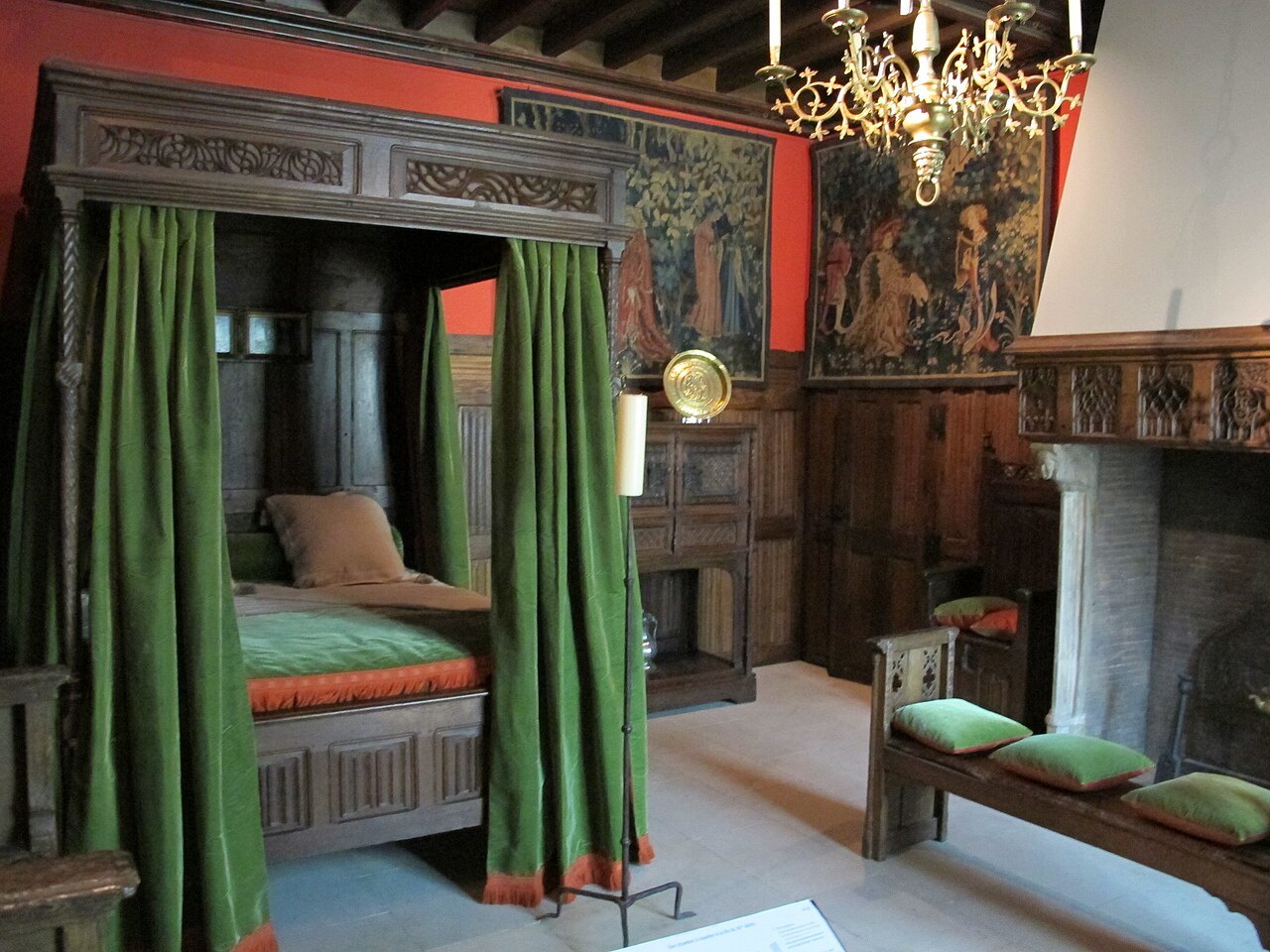 A carved wooden canopy bed with green curtains in a reconstructed medieval room at the Musée des Arts Décoratifs.