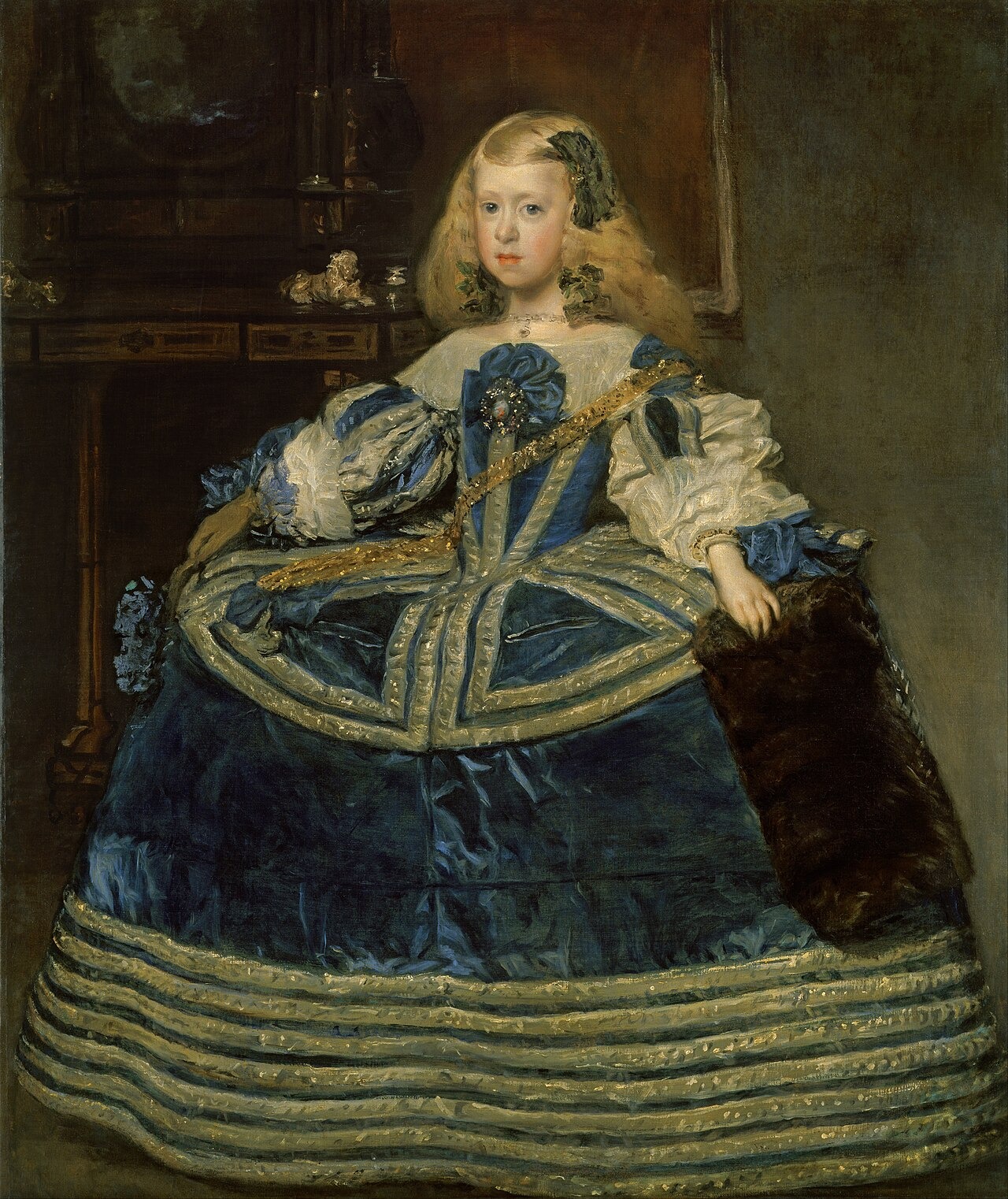 A young princess in a broad blue dress stands facing forward in a dark palace interior.