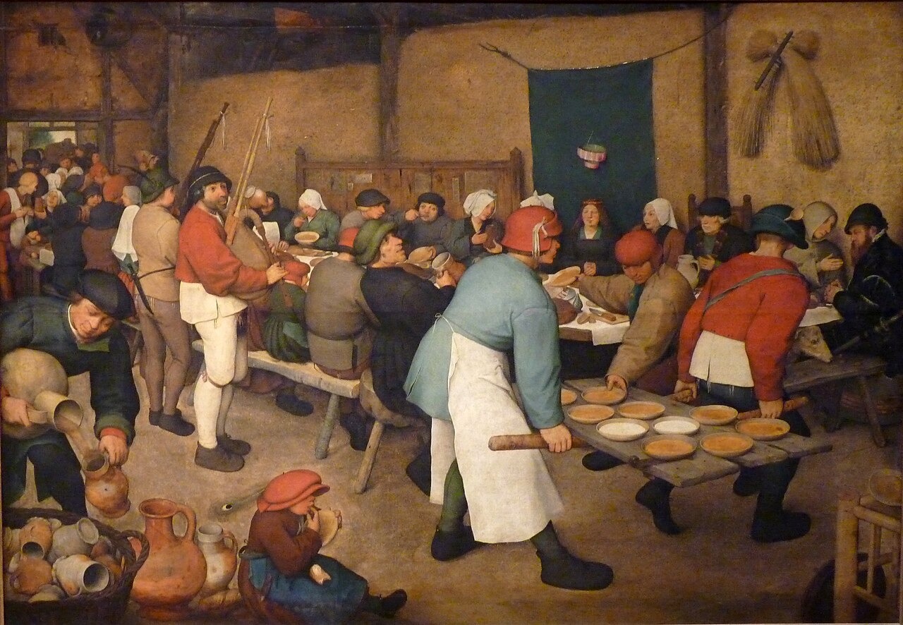 Guests crowd a barn around a long table while two men carry wooden boards laden with bowls.