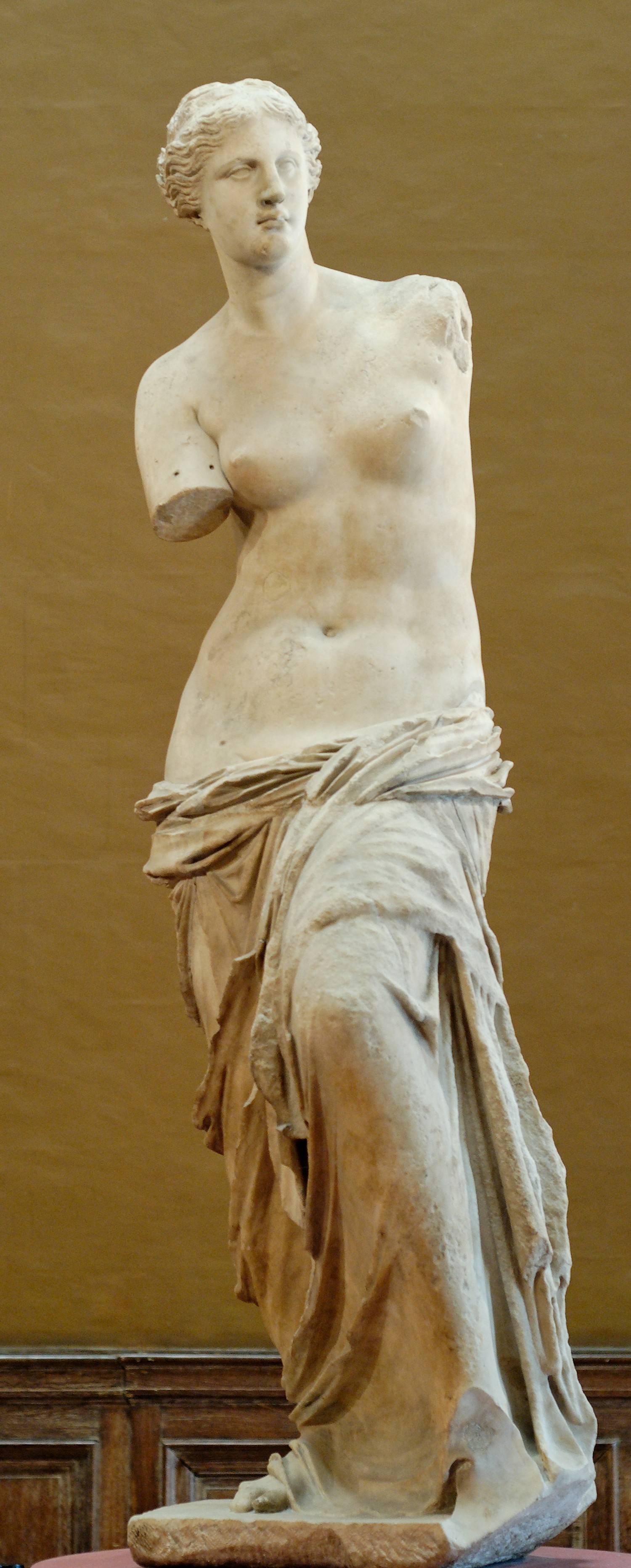 Venus de Milo’s white marble figure stands above its base in the Louvre collection.