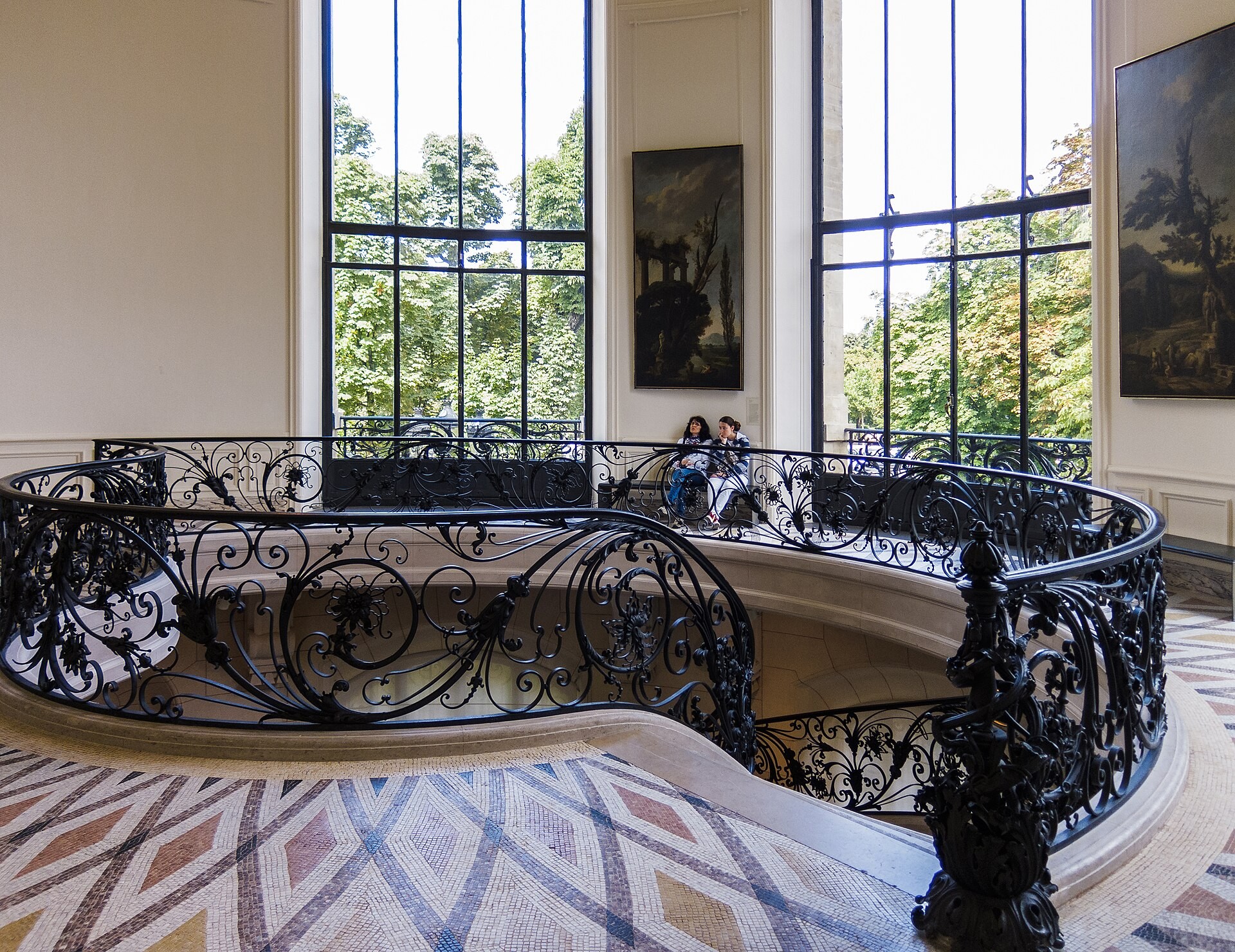 Black wrought-iron scrollwork curves around the Petit Palais staircase beneath tall windows.