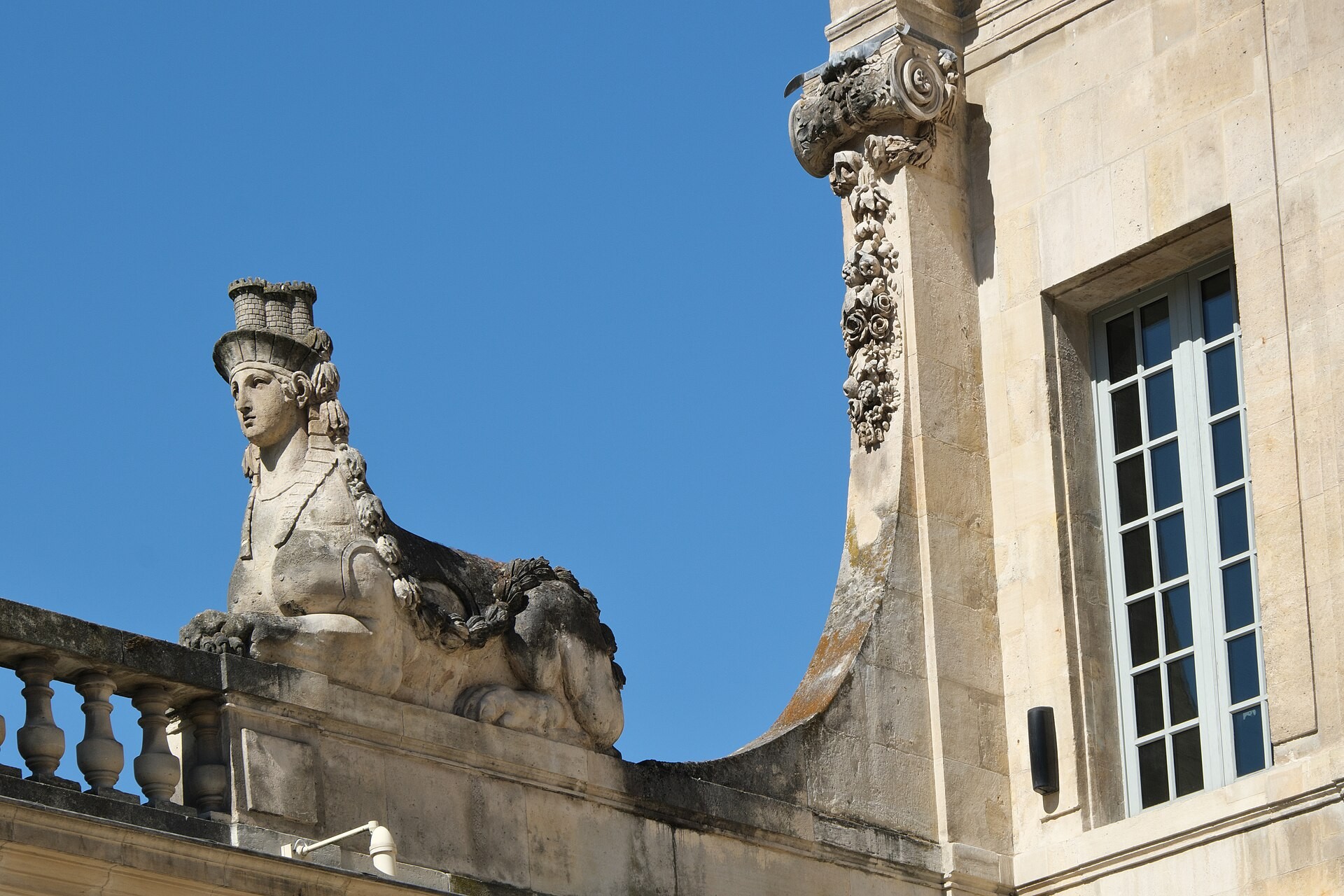 A carved stone sphinx rests on Hôtel Salé’s balustrade beside the decorated corner of its facade.