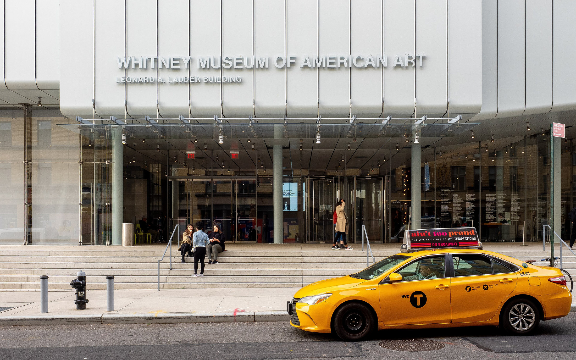 A yellow taxi passing the Whitney Museum’s glass entrance and name sign